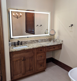 Bathroom vanity with wood cabinets, marble countertop, large backlit mirror, and wall sconce.