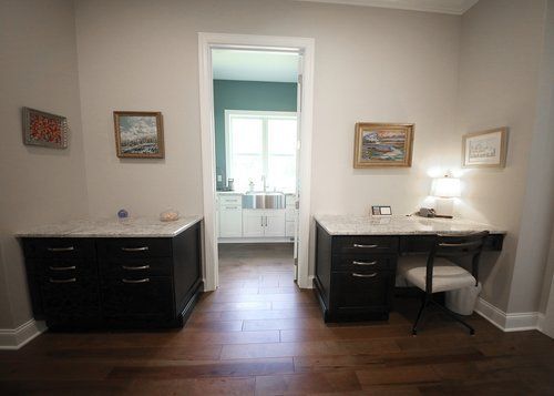 A home office with two dark desks, lamps, and a doorway to a bright bathroom beyond.