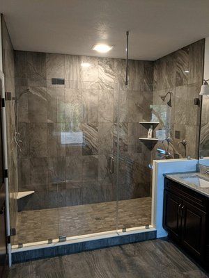 Walk-in shower with gray tile walls, glass enclosure, and blue-trimmed threshold beside a dark vanity.