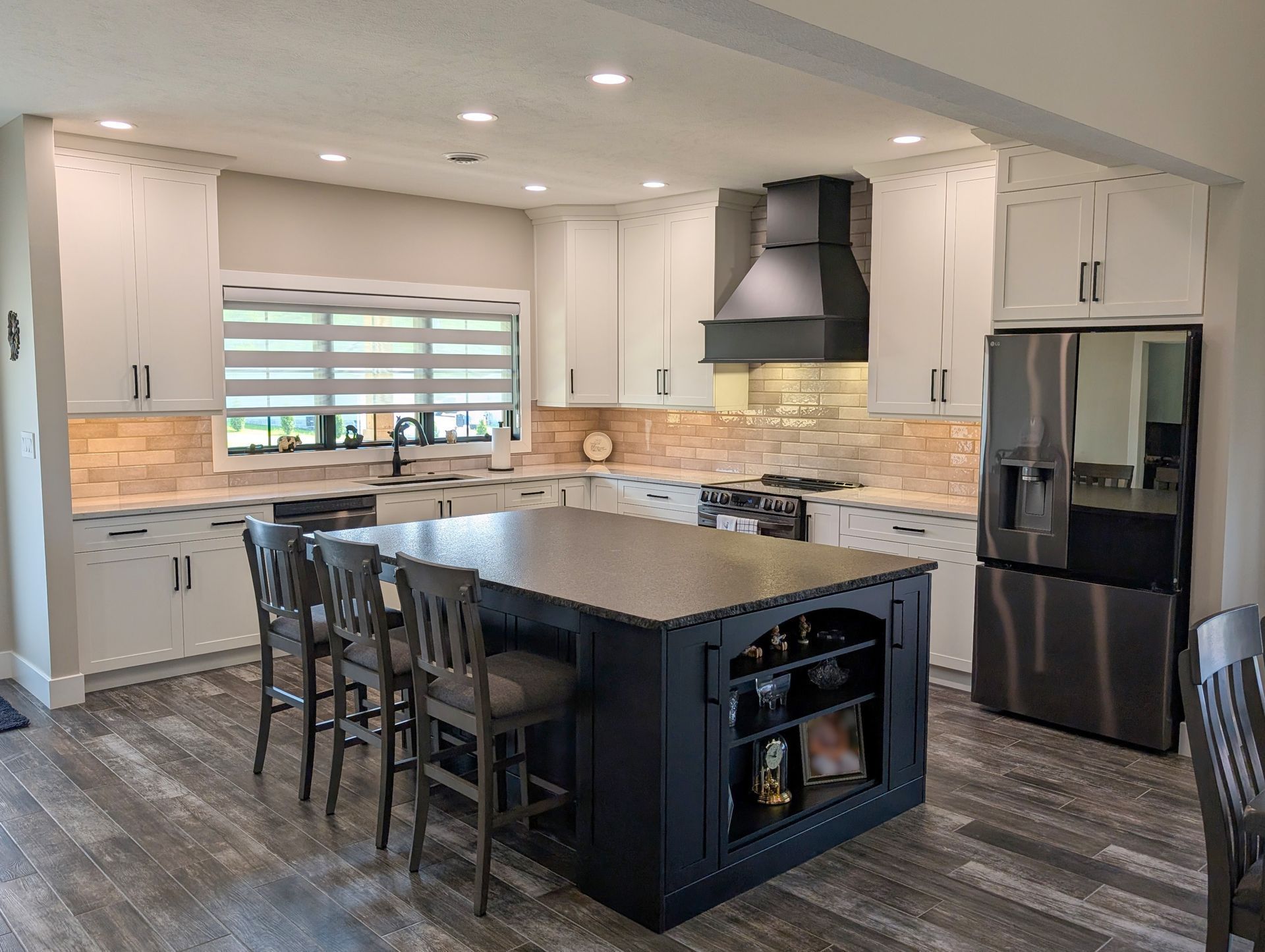 Modern kitchen with white cabinets, black island, stainless fridge, and bar stools beside a window.