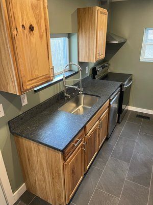 Compact kitchen with wood cabinets, black countertops, stainless sink, and dark tile floor.