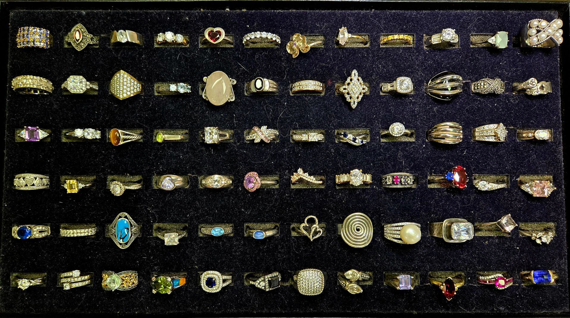 Rows of assorted rings and jewelry displayed on dark fabric, with gold, silver, and colorful stones