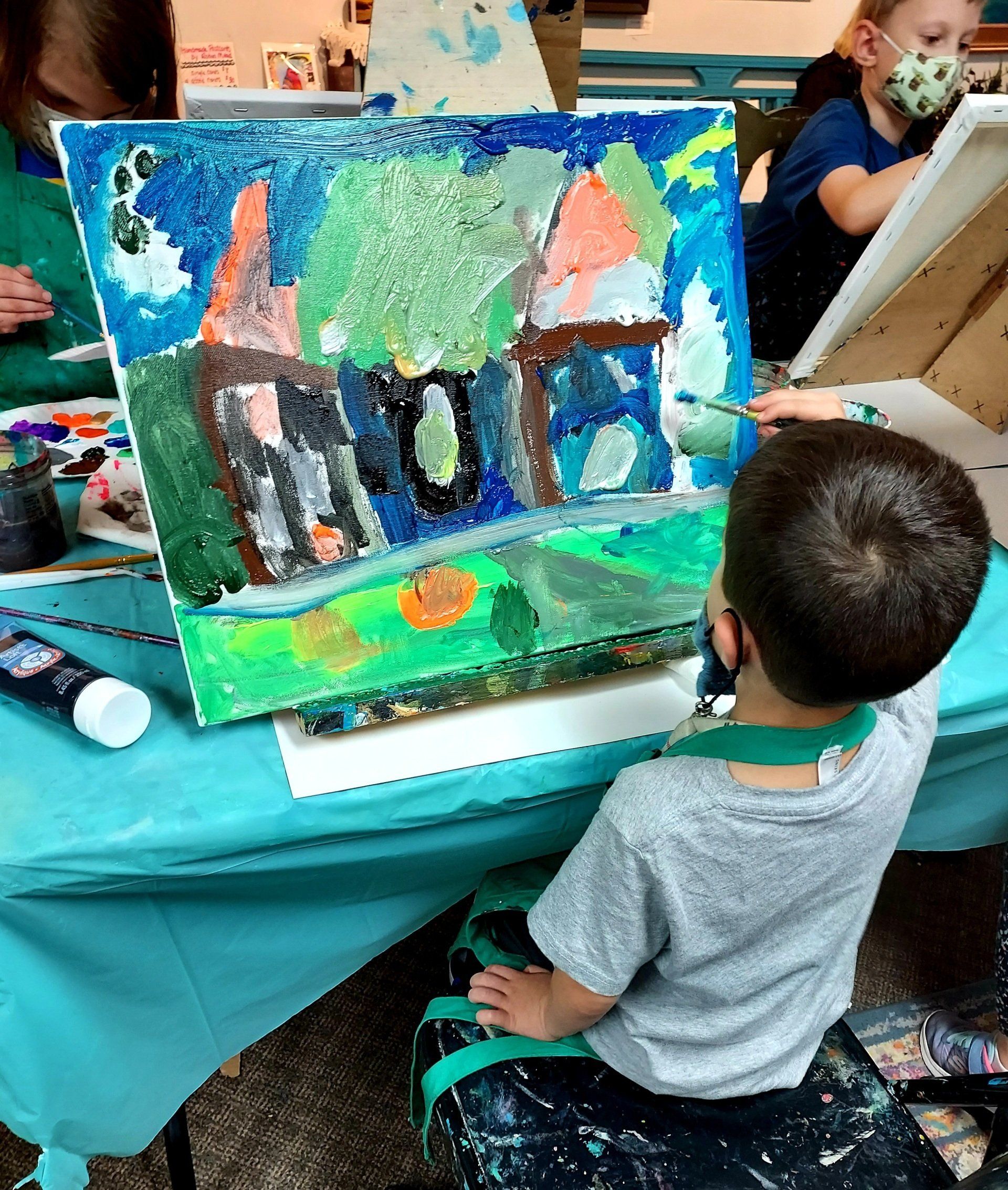 A young boy is sitting at a table holding a painting of a house.