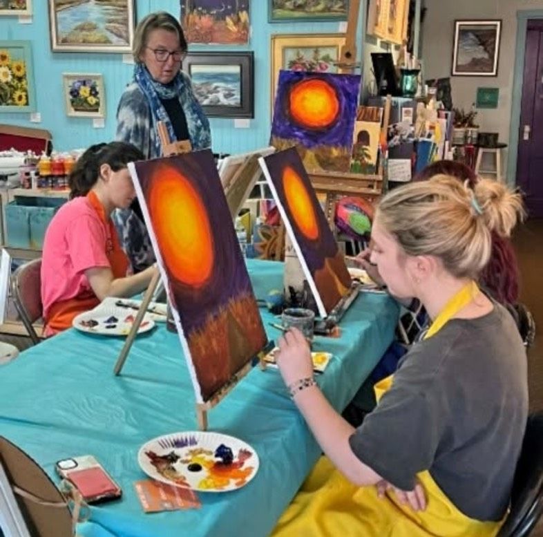 A woman sits at a table painting a picture of the sun