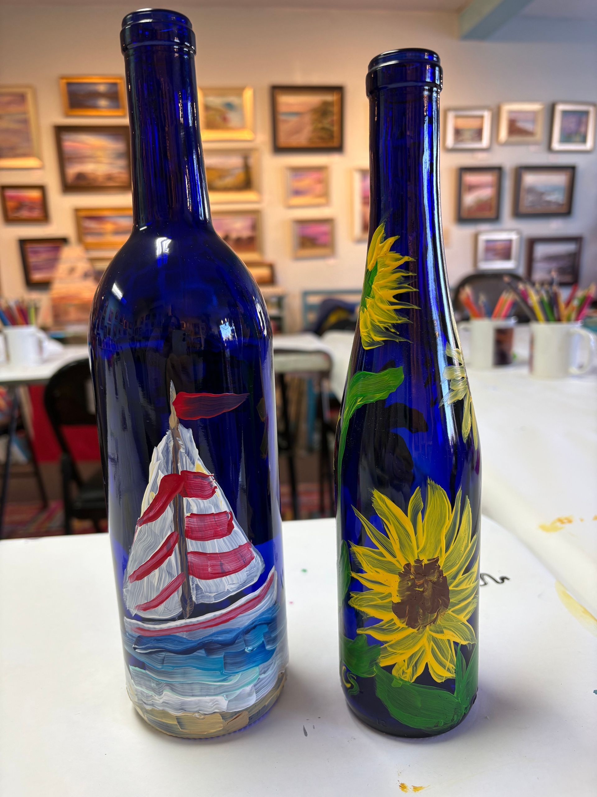 Two blue wine bottles with sailboats and sunflowers painted on them are sitting on a table.
