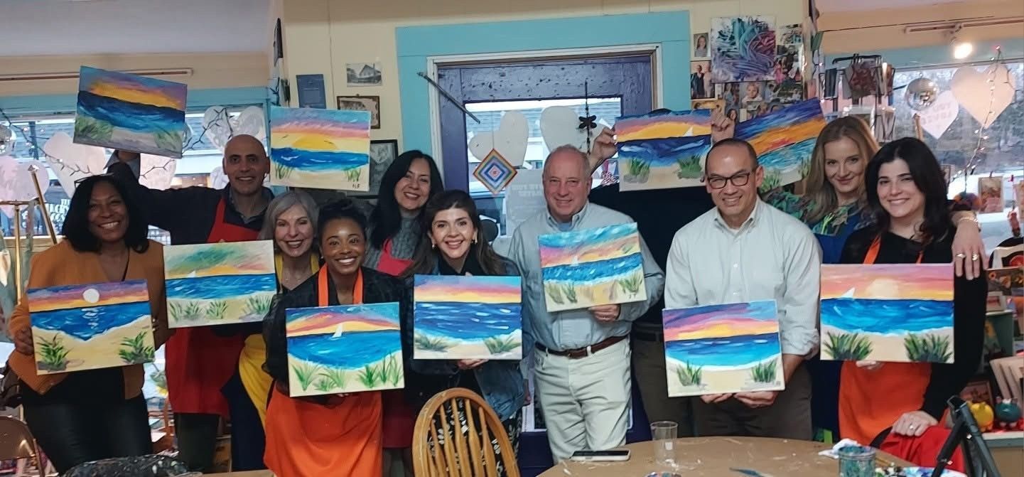 Group of people holding colorful landscape paintings in an art studio, smiling at the camera