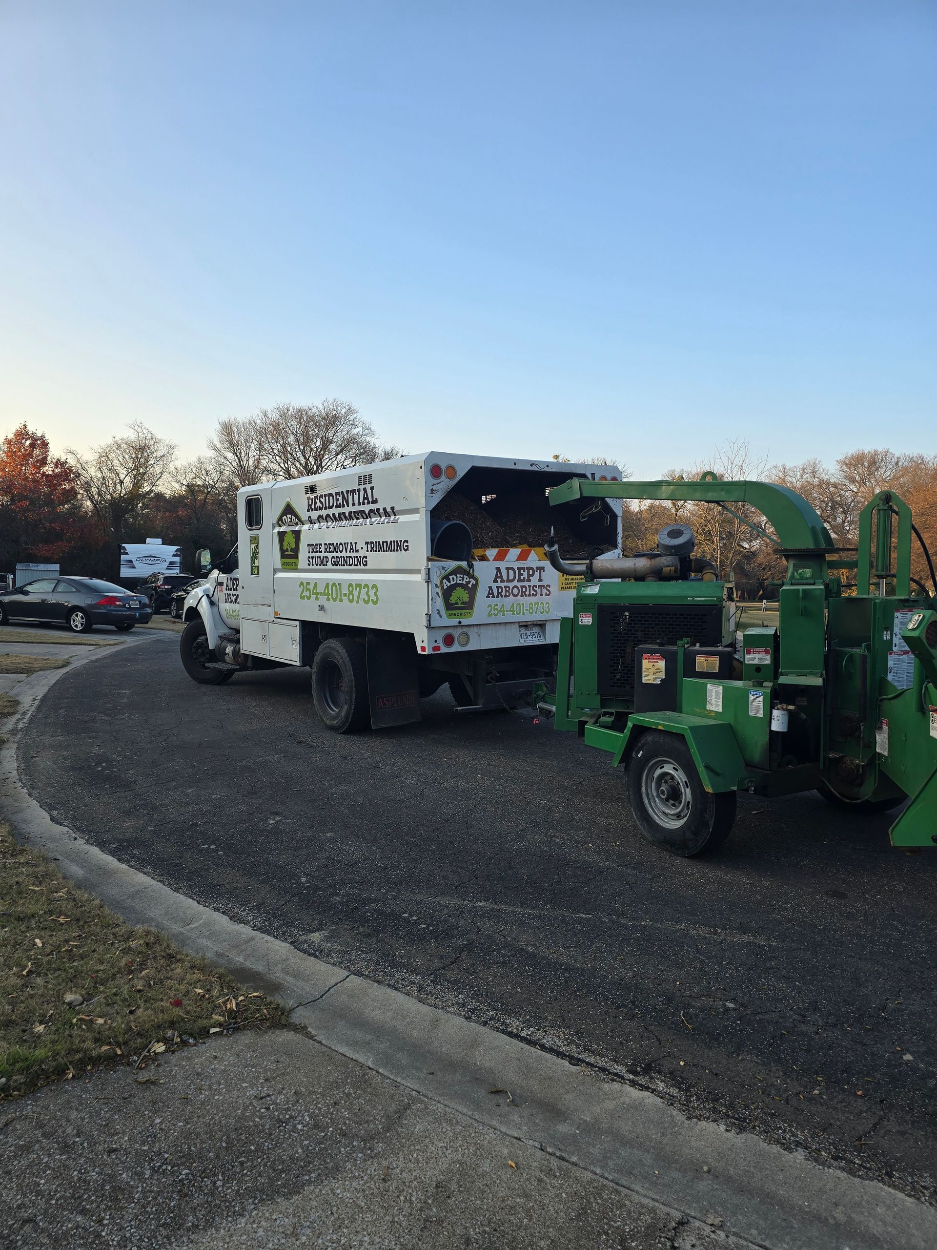 White utility truck and green tree chipper on a gravel road at dusk, with equipment parked beside them