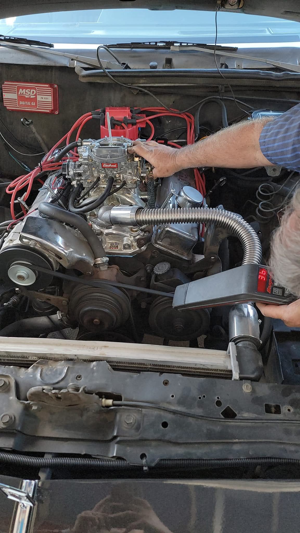 An individual works on a car engine, holding a timing light aimed at the front of the motor in an open engine bay.
