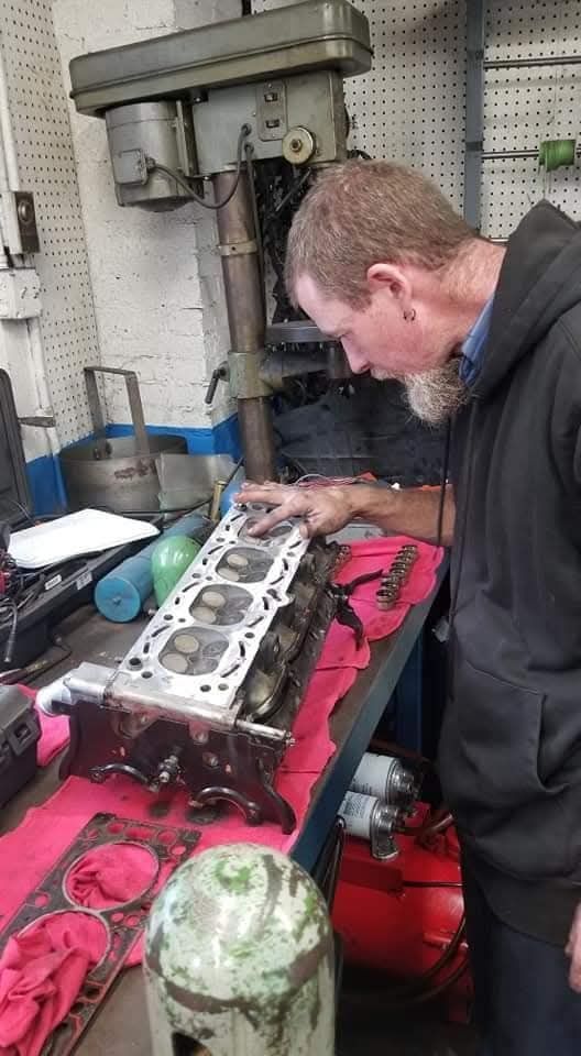 A person works on a metal engine cylinder head in a workshop, placing a gasket on top of the engine block.