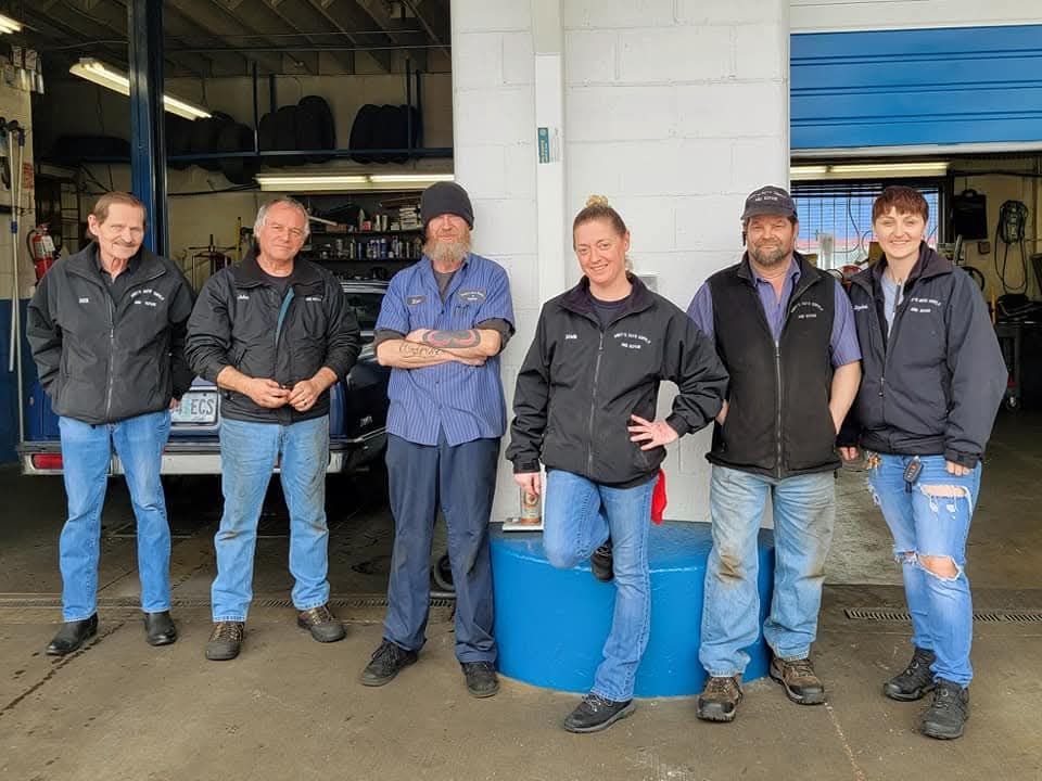 Six people in work uniforms stand in a row inside an auto repair shop, posing for a group photo.