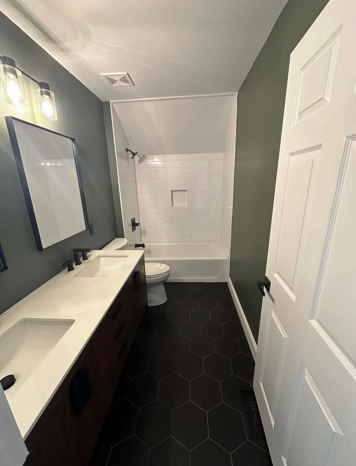 Narrow bathroom with dark hex tile floor, white vanity and door, mirror, and shower at the far end.
