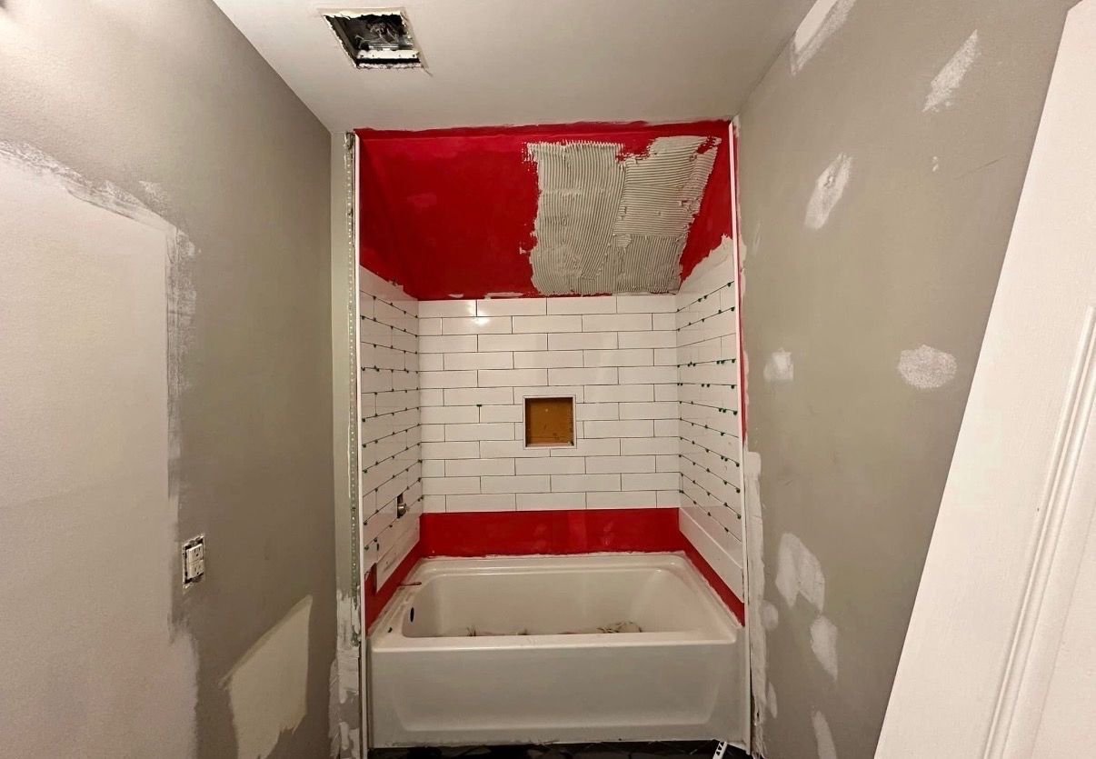 Bathroom shower alcove under renovation with red waterproofing, white tile, and a tub base.