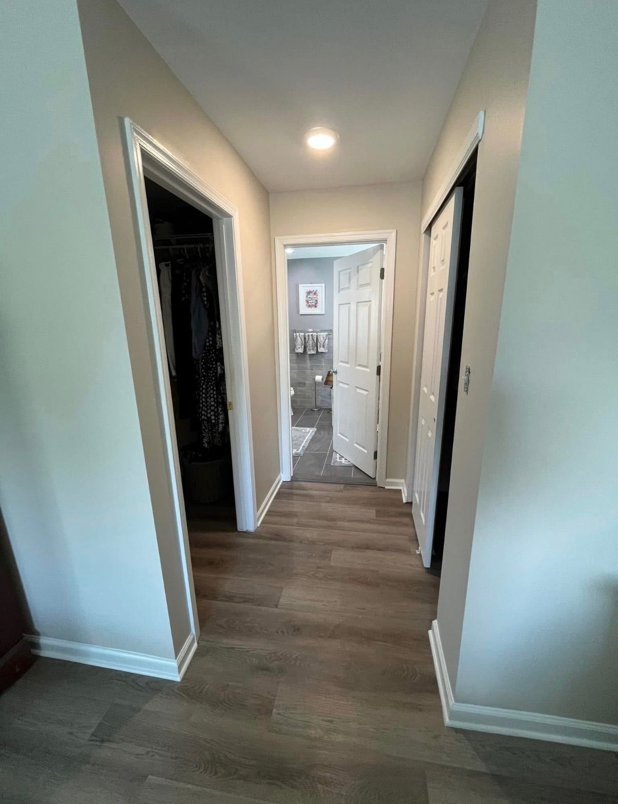 Narrow hallway with wood floors, white walls, and several open doorways leading to a bright room.