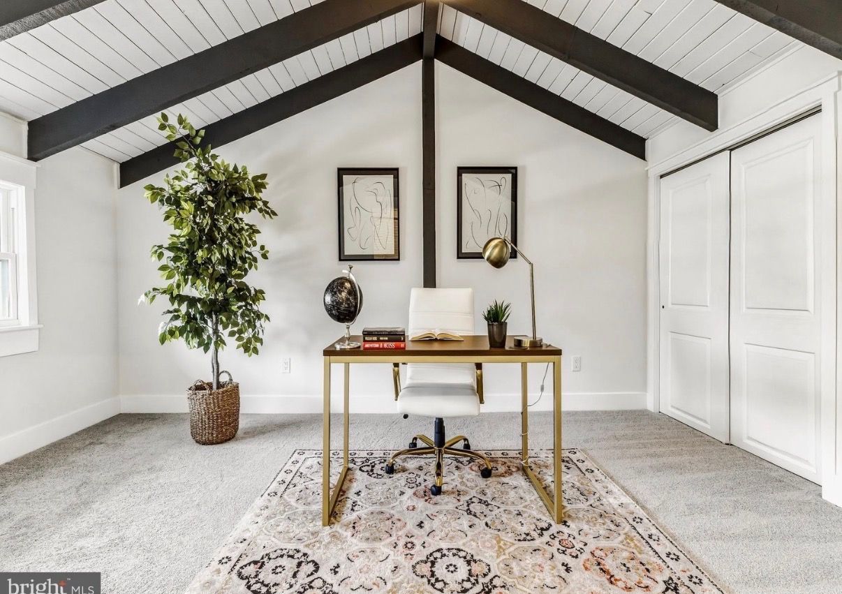 Minimal home office with white walls, gold desk, potted tree, and patterned rug under vaulted beams