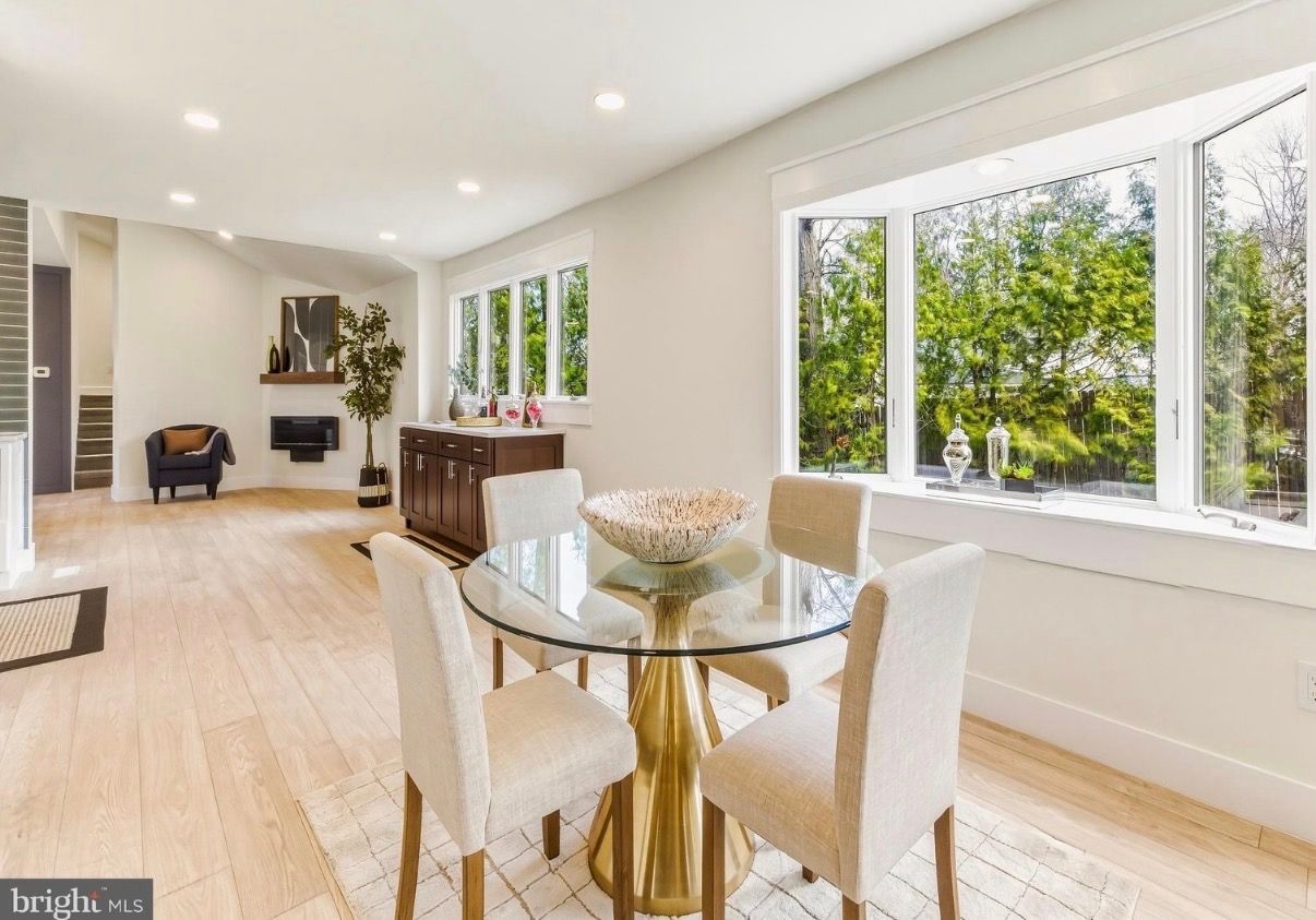 Bright dining room with glass table, cream chairs, and large windows overlooking green trees