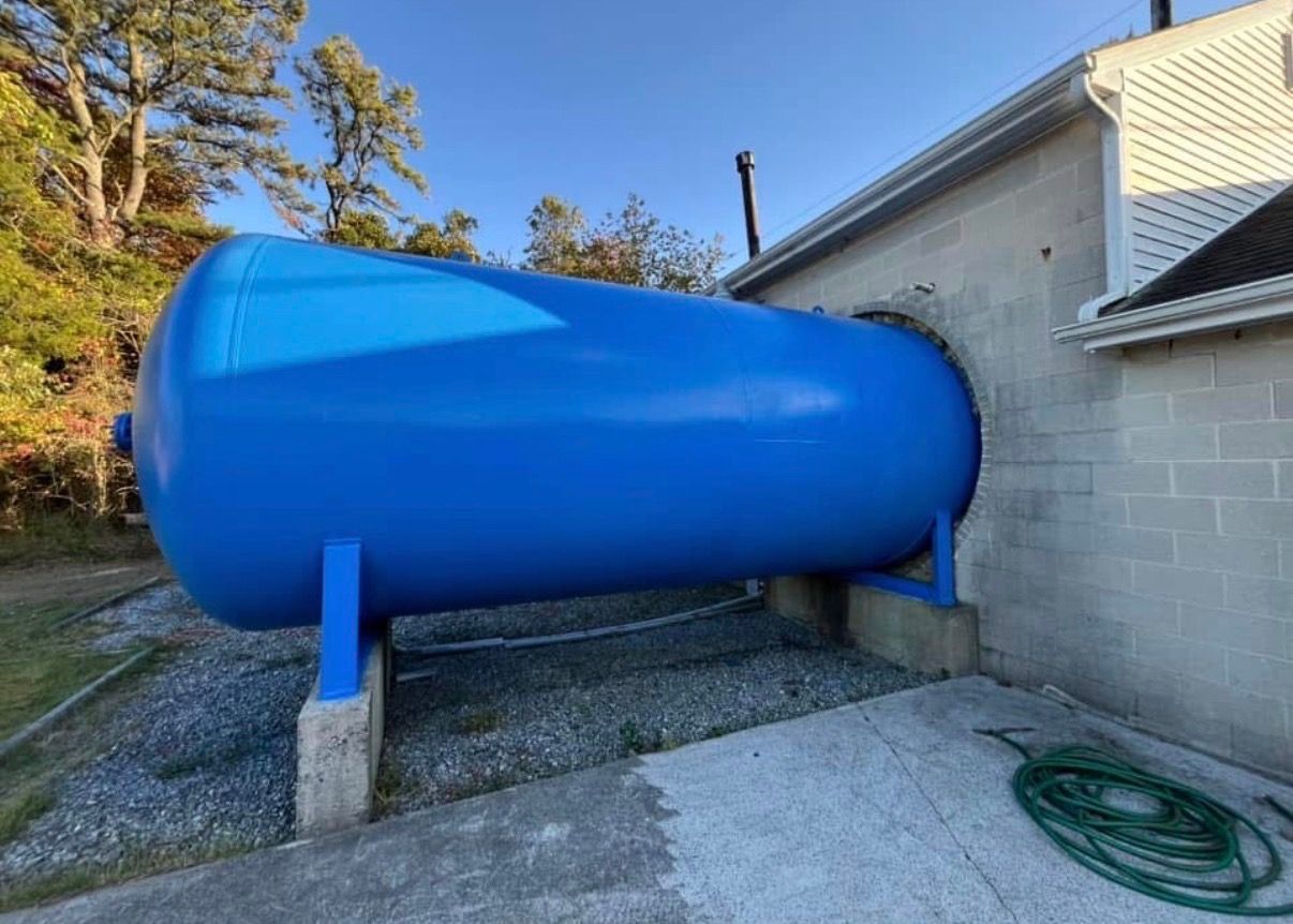 Blue horizontal storage tank beside a house on supports, with a hose on the ground