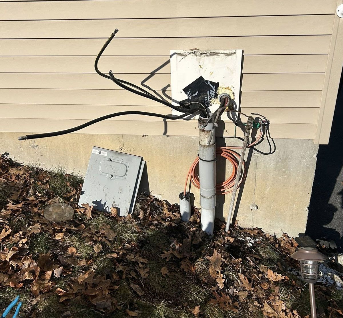 Damaged outdoor electrical meter box with exposed wires beside a house foundation and fallen leaves