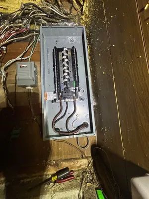 Open electrical breaker panel with exposed wiring in a wooden wall cavity, tools below.