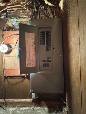 Wall-mounted electrical breaker panel in a dim basement with exposed wires and wood framing.