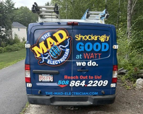 Blue electric company van with “The Mad Electrician” logo and contact info on the back, parked outdoors.