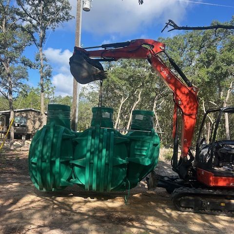 A green septic tank lifted by an orange excavator in a wooded area