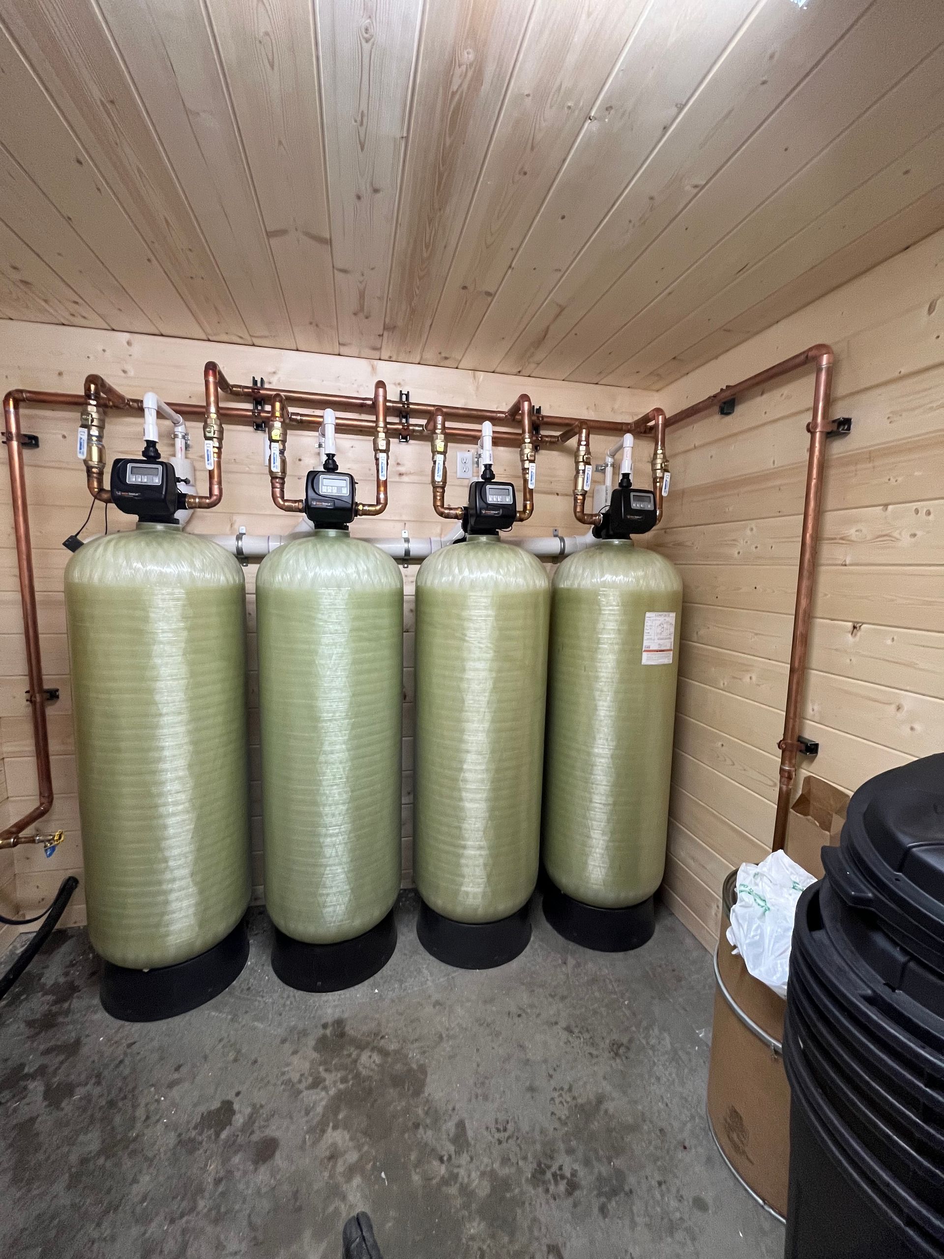 Four cylindrical water filtration tanks with copper pipes in a small wooden room.