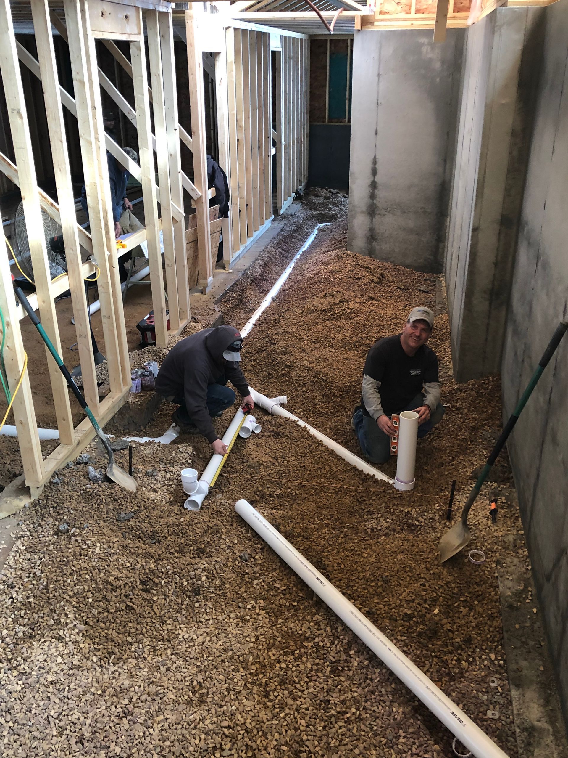Two workers installing PVC pipes in a trench filled with wood chips, indoors, near unfinished walls.