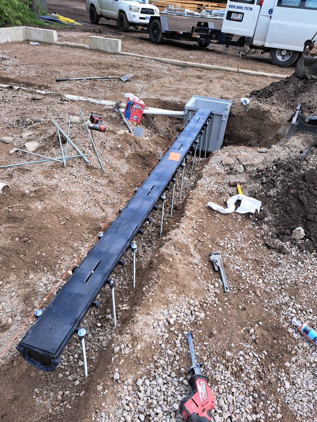 Construction site with a long, dark utility box in a trench. Metal rods and tools are scattered around.