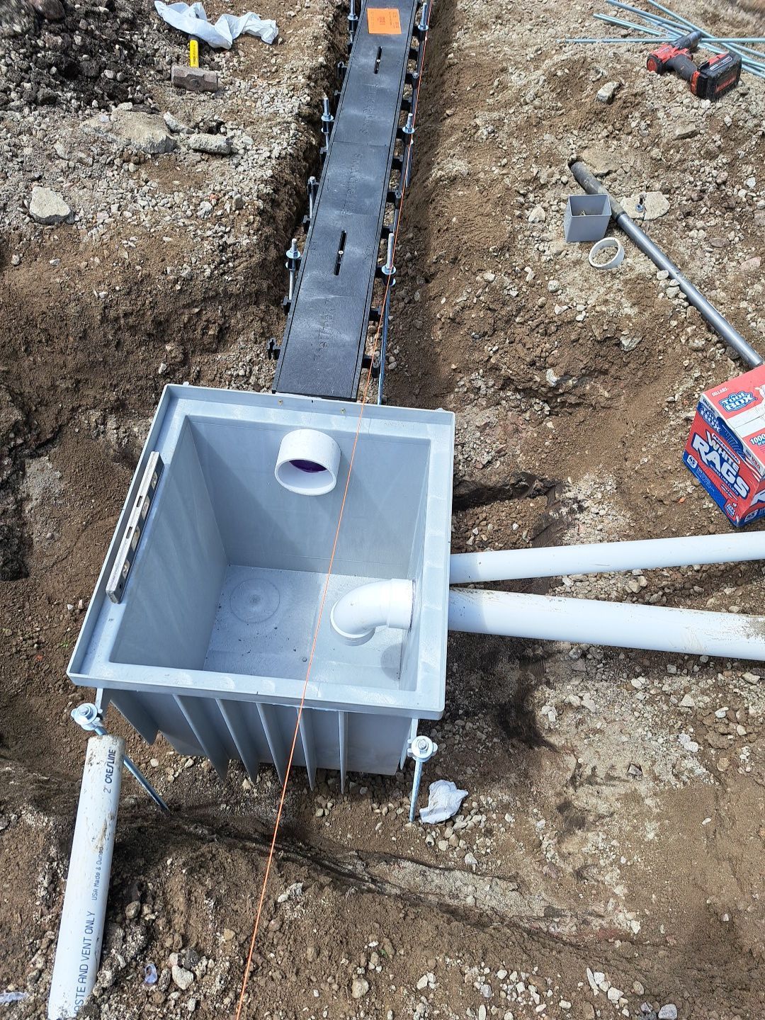Gray utility box with connected pipes, installed in a trench. A long, gray conduit runs along the trench.