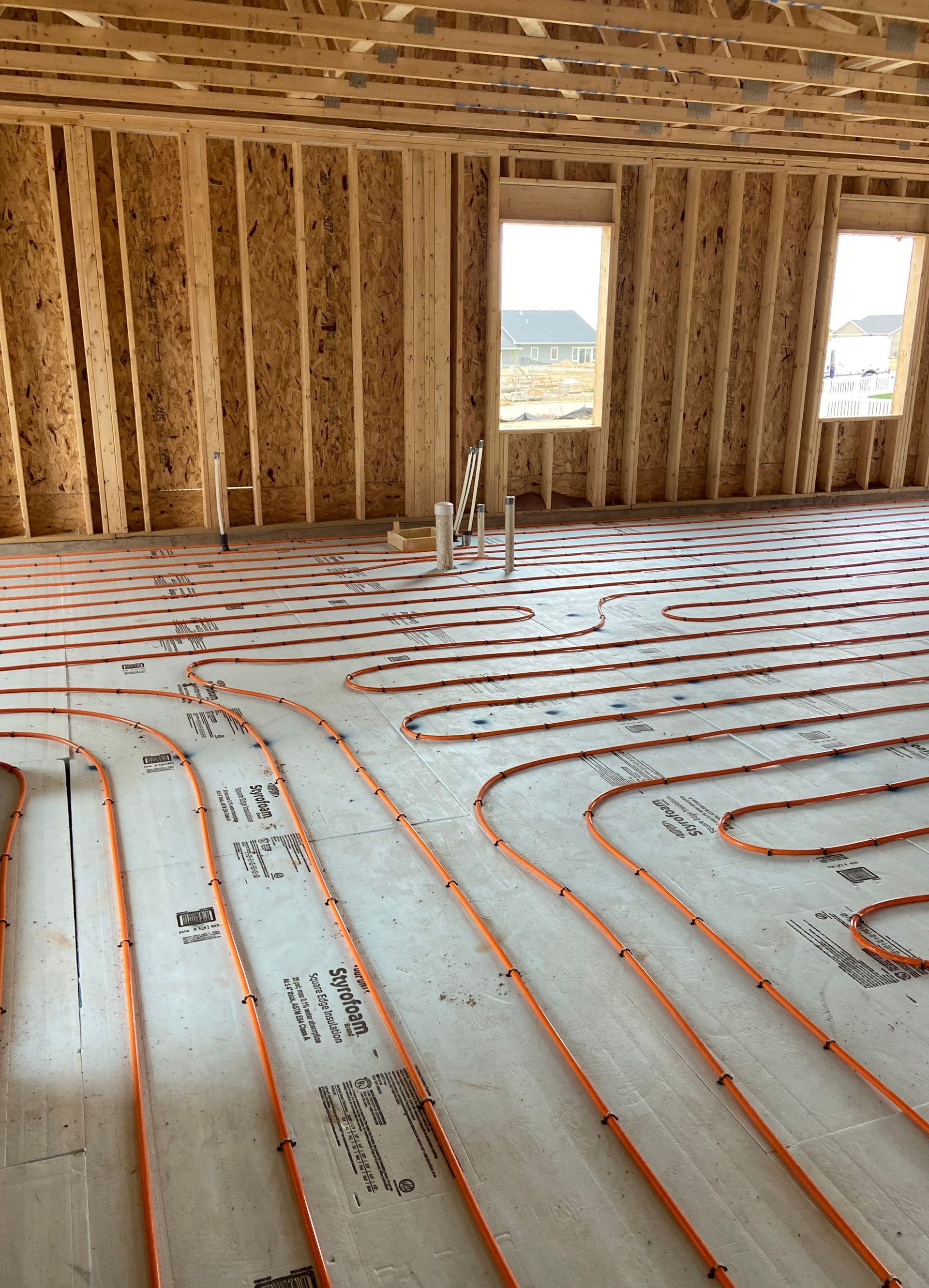 Construction interior showing orange radiant heating tubes laid on concrete floor. Wooden frame walls and ceiling with windows.