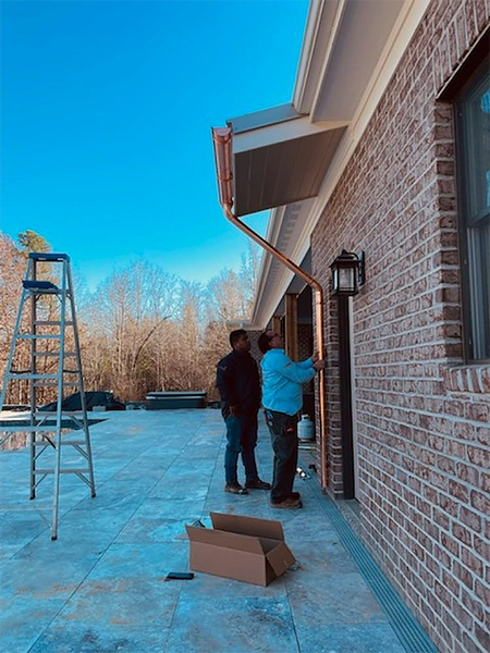 Two workers installing copper gutter on a brick house; ladder and cardboard box on a patio.