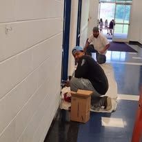 Two men painting a wall indoors. One kneels, the other stands. White wall, blue door.
