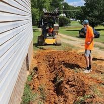 Man watches a skid steer dig a trench next to a house with white siding.