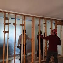 Two people in a room being renovated; one is working, the other is wearing a red hat. Wooden frame visible.