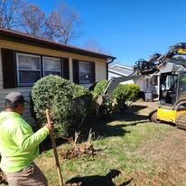 Man in green jacket watches excavator removing bushes next to a house.