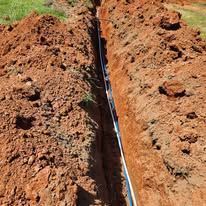 Trench dug in red soil, containing a white pipe.
