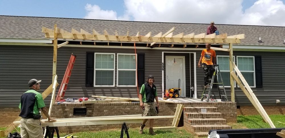 Workers constructing a pergola on a house porch. Wooden beams and tools are visible.