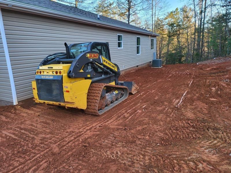 Yellow skid steer on red dirt next to a light-colored house.