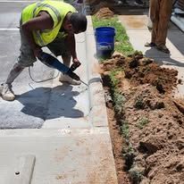 Person in safety vest using a jackhammer on concrete curb next to dirt and grass.