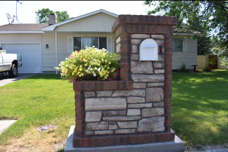 Stone and brick mailbox with flowers in front of a light-colored house.