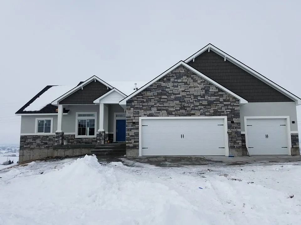 House with stone and grey siding, two-car garage, snow-covered yard, cloudy sky.