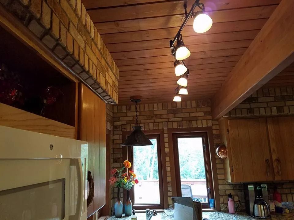 Kitchen with brick walls and wooden ceiling, track lighting, cabinets, microwave, and windows with greenery.