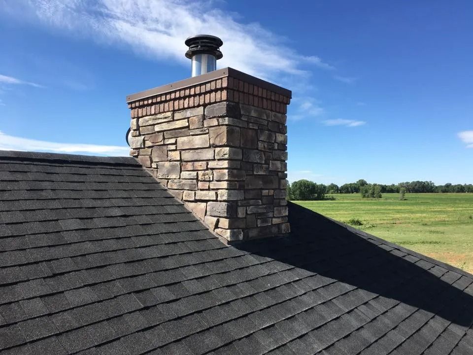 Stone chimney on a dark shingle roof against a blue sky, overlooking a green field.