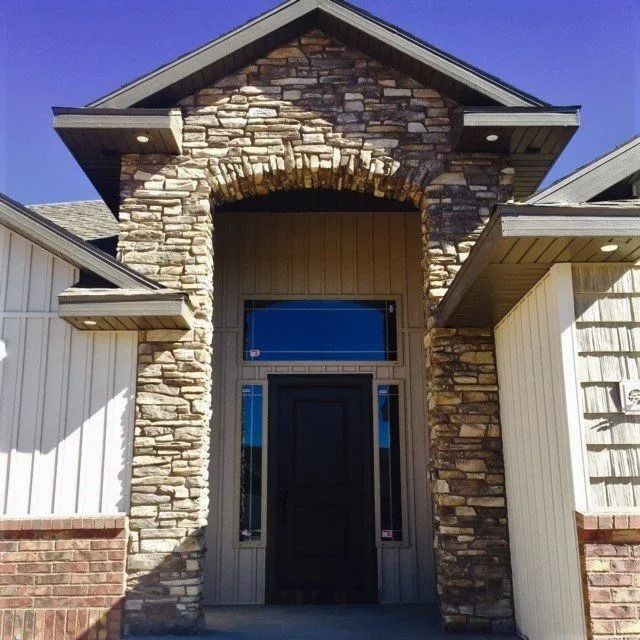 Stone-covered entryway of a modern house; black door, sidelights, and a transom window.