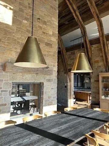 Two brass cone pendant lights hang over a dark rug-covered dining table. Stone walls and wooden beams in the room.