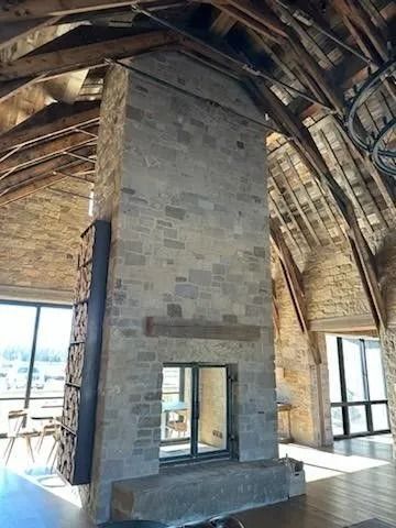 Stone fireplace in a vaulted room with exposed wooden beams, firewood rack, and large windows.