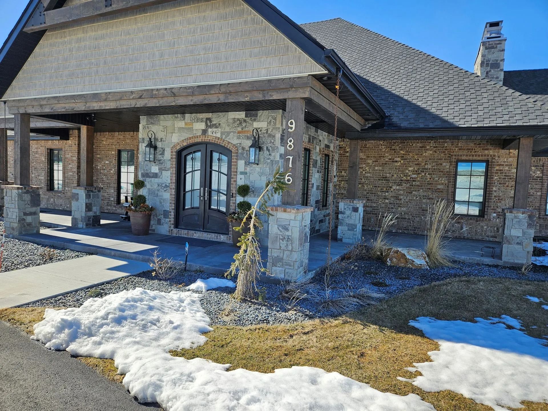 Stone and brick home exterior with snow on the lawn. A covered porch leads to double doors with the number 9876.