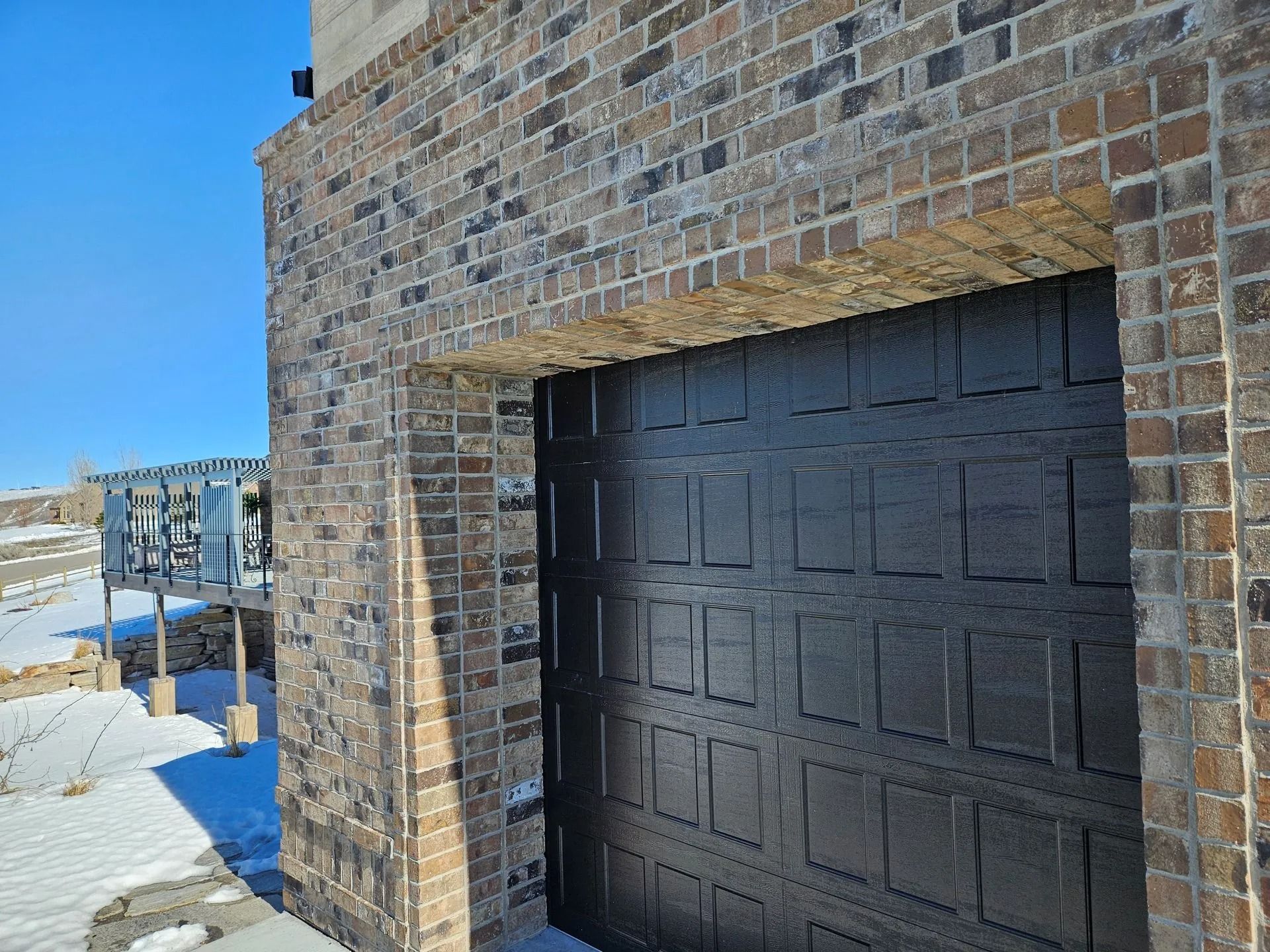 Black garage door framed by a brick wall, snowy landscape, and clear blue sky.
