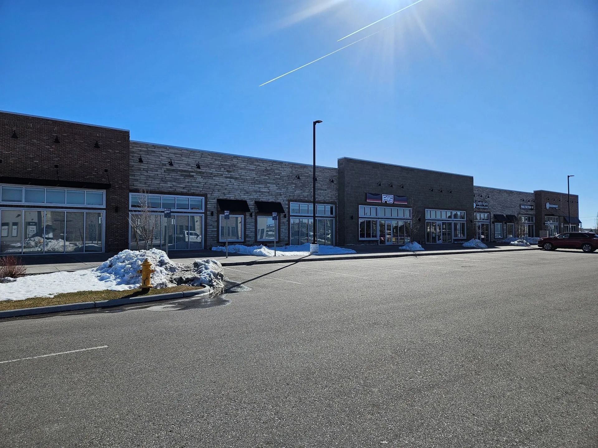 Long, brick building with multiple storefronts and a parking lot. Snow on the ground under a bright blue sky.