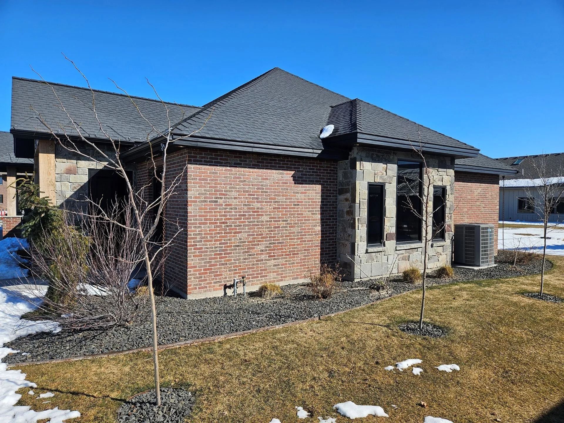 Brick and stone house with dark gray roof, dry grass, some snow, and blue sky.